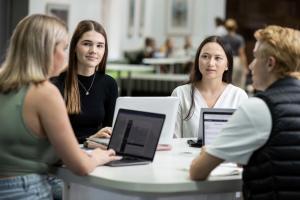 Students sit at a round desk facing each other with laptops in front of them