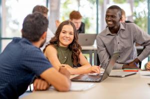 Three students sit in class with books and a laptop in front of them 